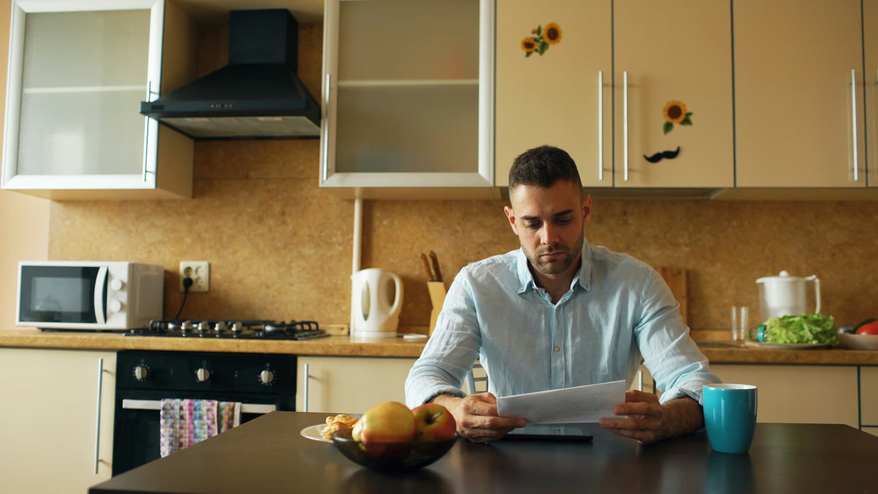 Un homme assis à la table de sa cuisine lit attentivement un courrier, une tasse posée à côté de lui et une tablette sous la feuille.