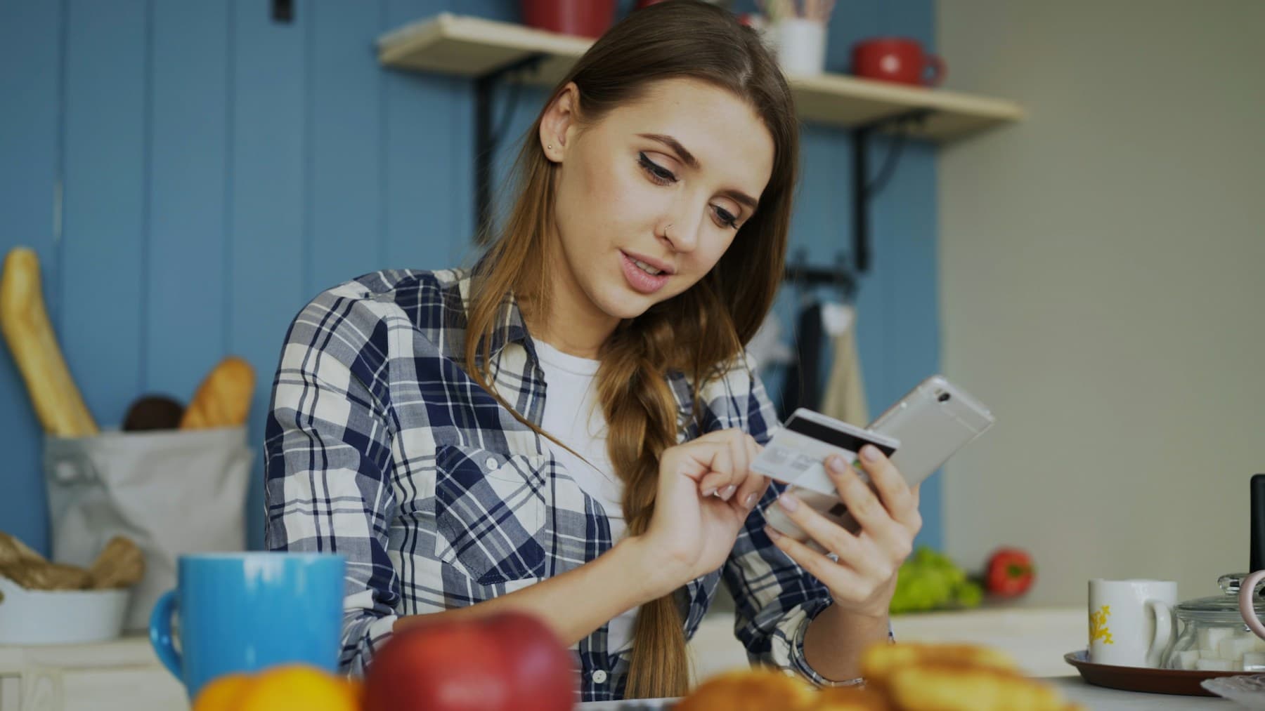 Une jeune femme assise dans sa cuisine saisit quelque chose sur son téléphone en tenant une carte bancaire dans la même main. Sur la table, des fruits et une tasse.