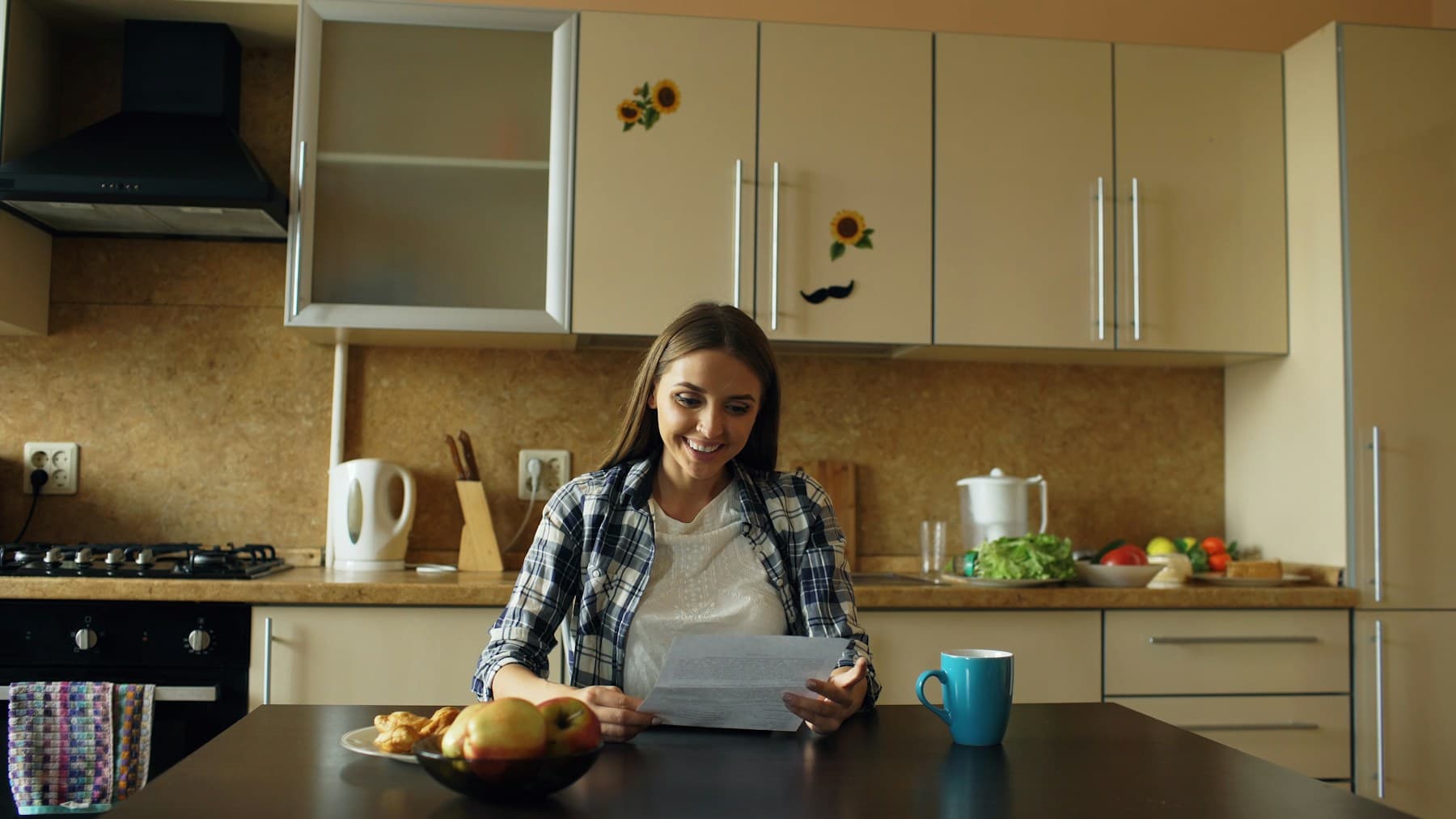 Une femme assise à la table de sa cuisine lit une feuille de papier qu'elle tient à deux mains, en souriant. Devant elle, une tasse bleue et une coupe de fruits.